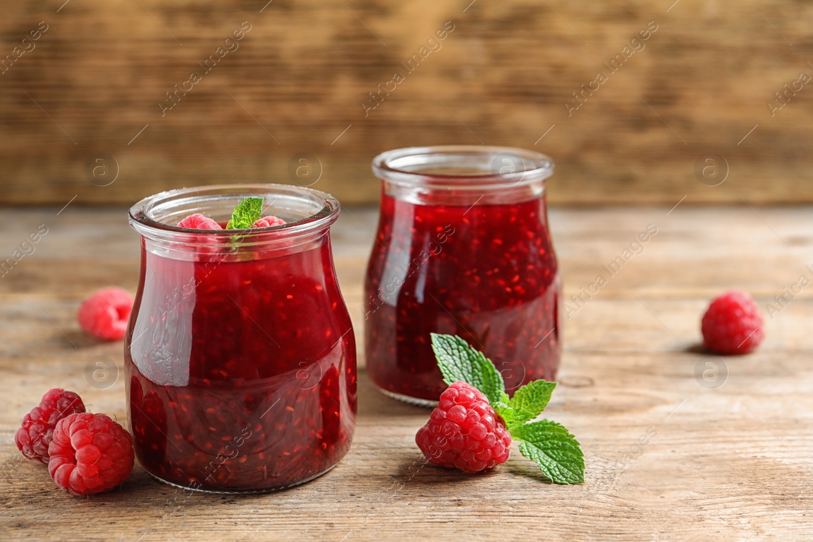 Photo of Glass jars of sweet jam with ripe raspberries and mint on wooden table. Space for text