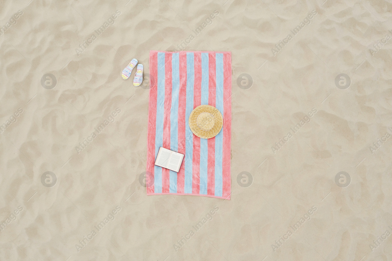 Image of Striped beach towel, book, straw hat and flip flops on sand, aerial view