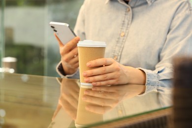 Photo of Woman holding takeaway paper cup and smartphone at table, closeup. Coffee to go