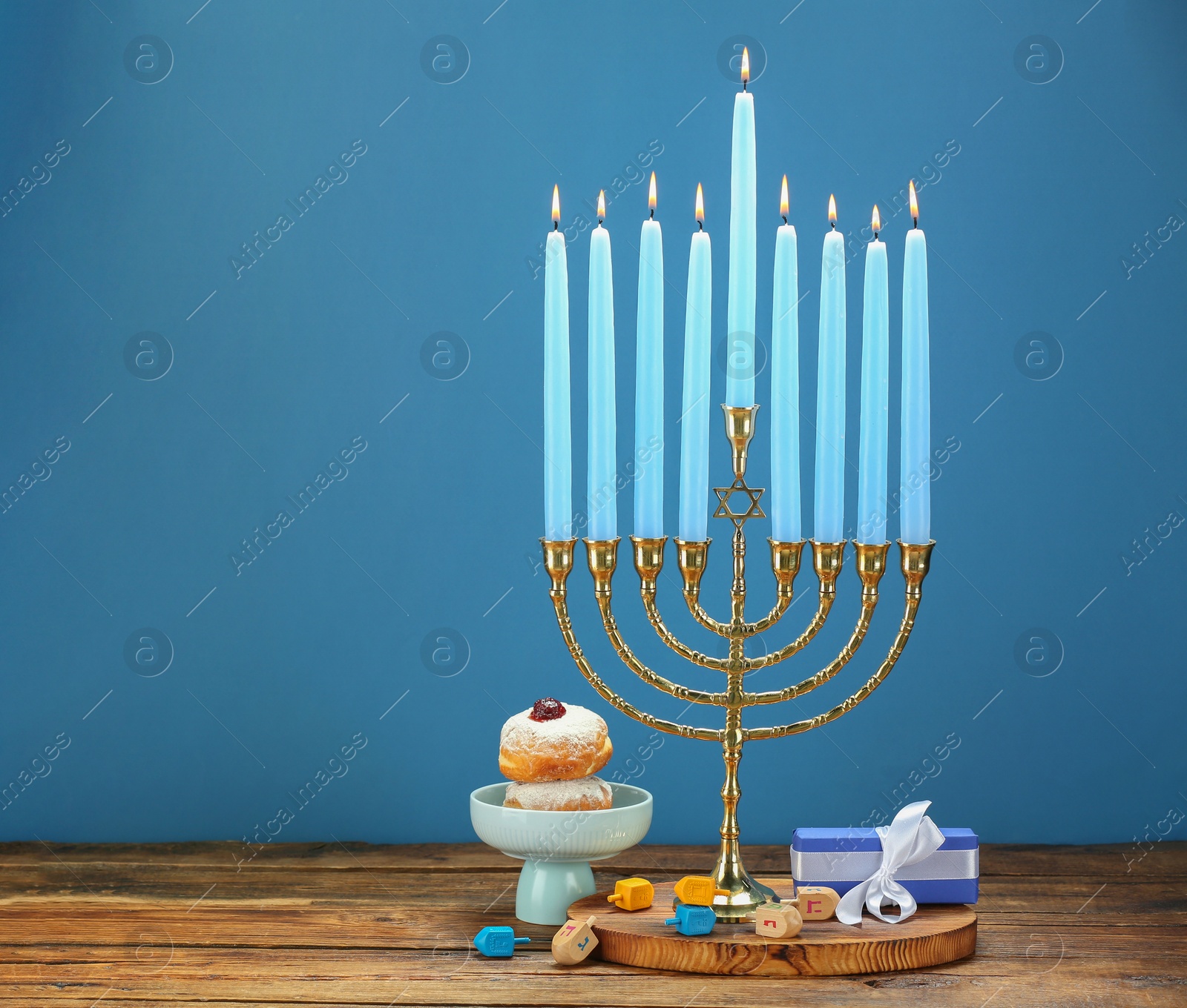 Photo of Hanukkah celebration. Menorah with burning candles, dreidels, donuts and gift box on wooden table against blue background, space for text
