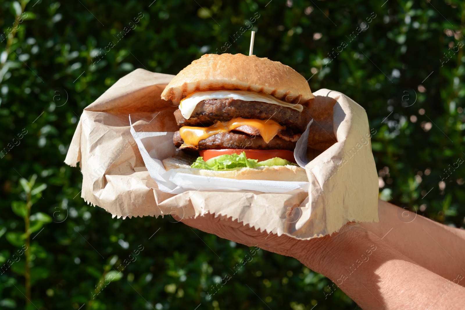 Photo of Woman holding delicious burger in paper wrap outdoors, closeup