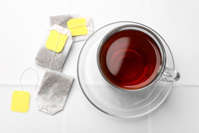 Tea bags and cup of aromatic drink on white tiled table, top view