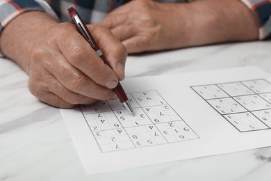 Photo of Senior man solving sudoku puzzle at white marble table, closeup