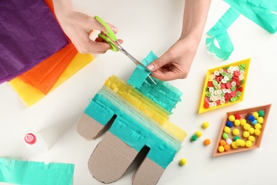 Photo of Woman cutting green paper for decorating cardboard cactus at white table, top view. Pinata DIY