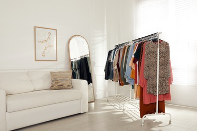 Photo of Rack with stylish clothes near sofa and mirror indoors. Fast fashion