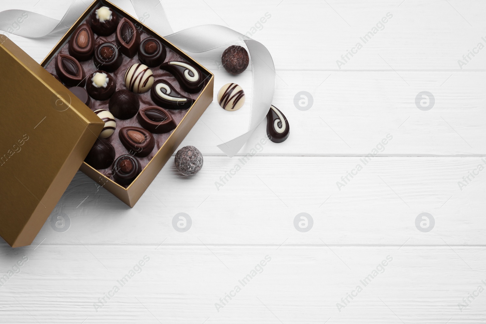 Photo of Open box of delicious chocolate candies and color ribbon on white wooden table, flat lay. Space for text