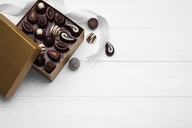 Open box of delicious chocolate candies and color ribbon on white wooden table, flat lay. Space for text