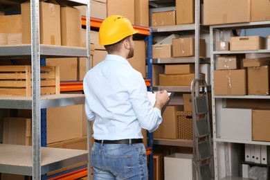 Photo of Young man with clipboard near rack of cardboard boxes at warehouse, back view