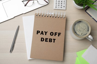 Image of Notebook with phrase Pay Off Debt, pen and cup of coffee on wooden table, flat lay