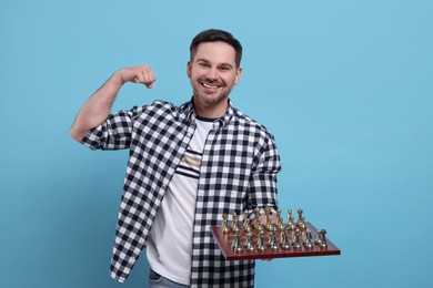 Photo of Smiling man holding chessboard with game pieces and showing bicep on light blue background