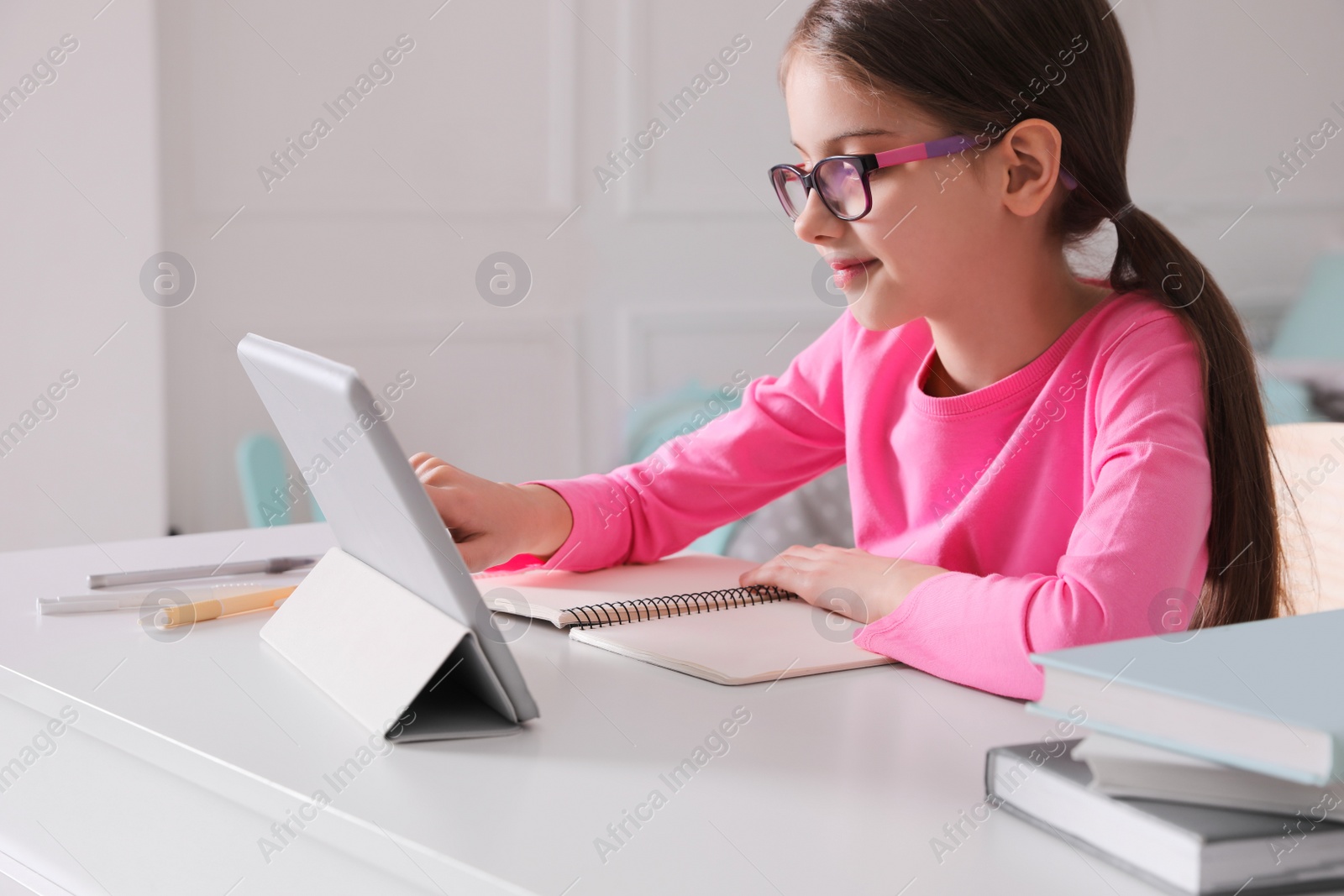 Photo of Little girl doing homework with tablet at table in room