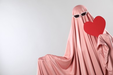 Photo of Cute ghost. Woman in pink sheet with sunglasses and red heart on light grey background, space for text