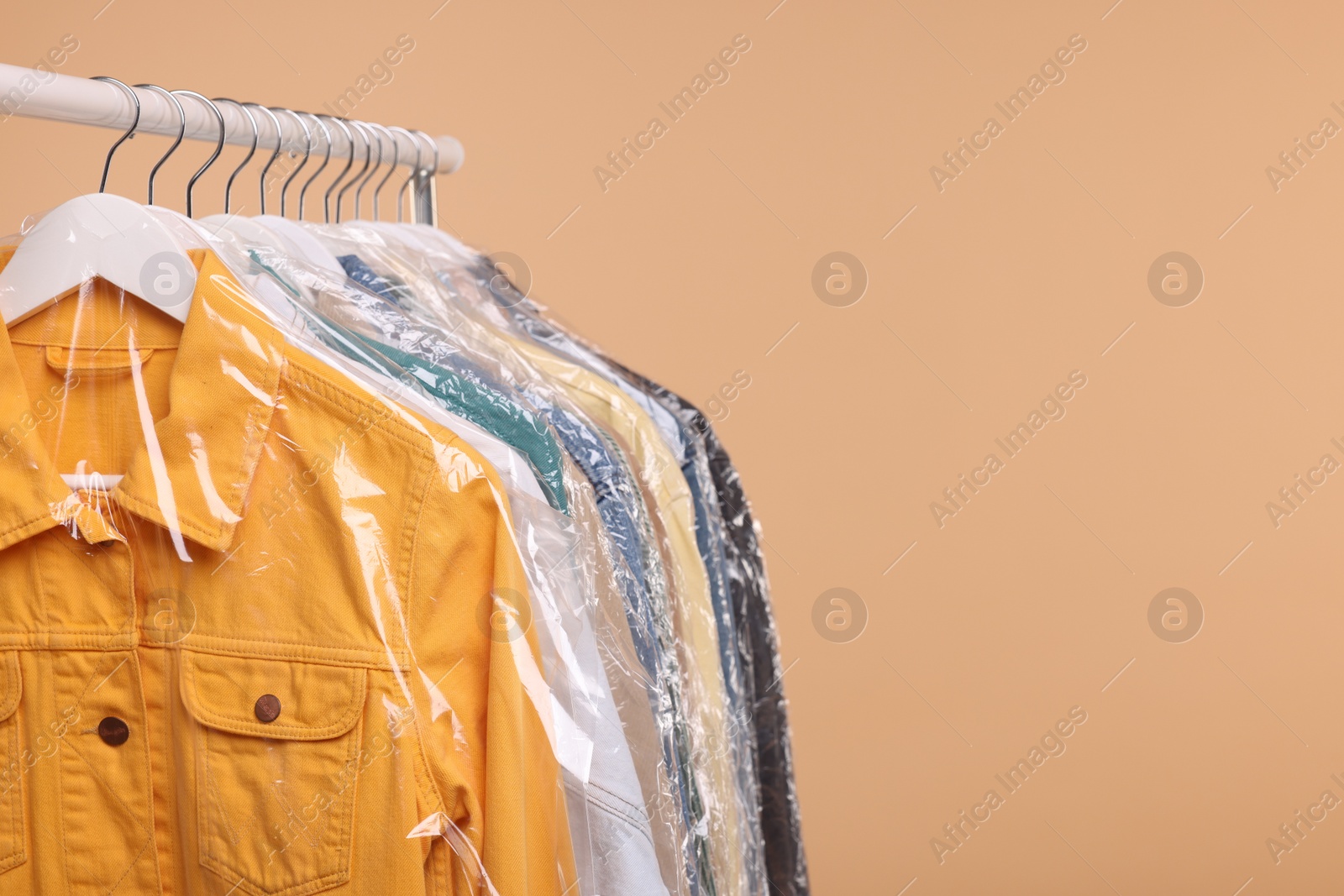 Photo of Dry-cleaning service. Many different clothes in plastic bags hanging on rack against beige background, space for text