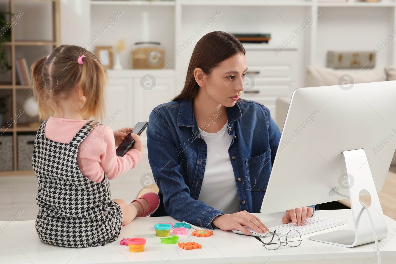 Photo of Woman working remotely at home. Woman using computer while her daughter playing with phone. Child sitting on desk