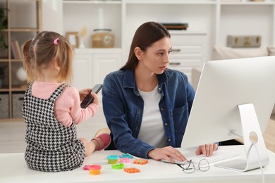 Photo of Woman working remotely at home. Woman using computer while her daughter playing with phone. Child sitting on desk