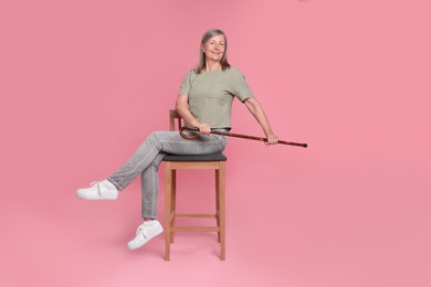 Senior woman with walking cane sitting on chair against pink background