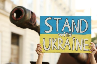 Photo of Woman holding poster in colors of national flag with words Stand with Ukraine near broken tank on city street, closeup