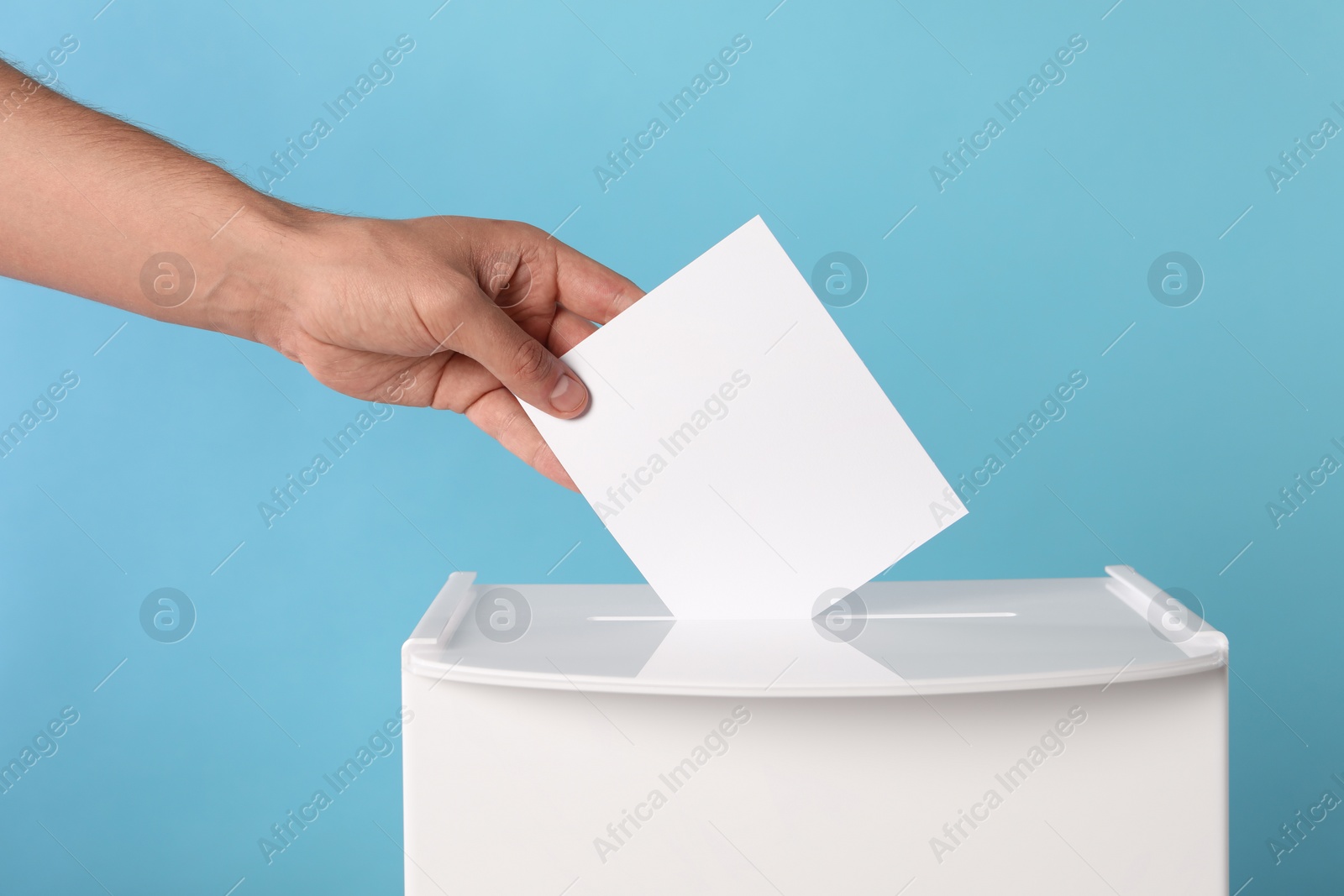 Photo of Man putting his vote into ballot box on light blue background, closeup