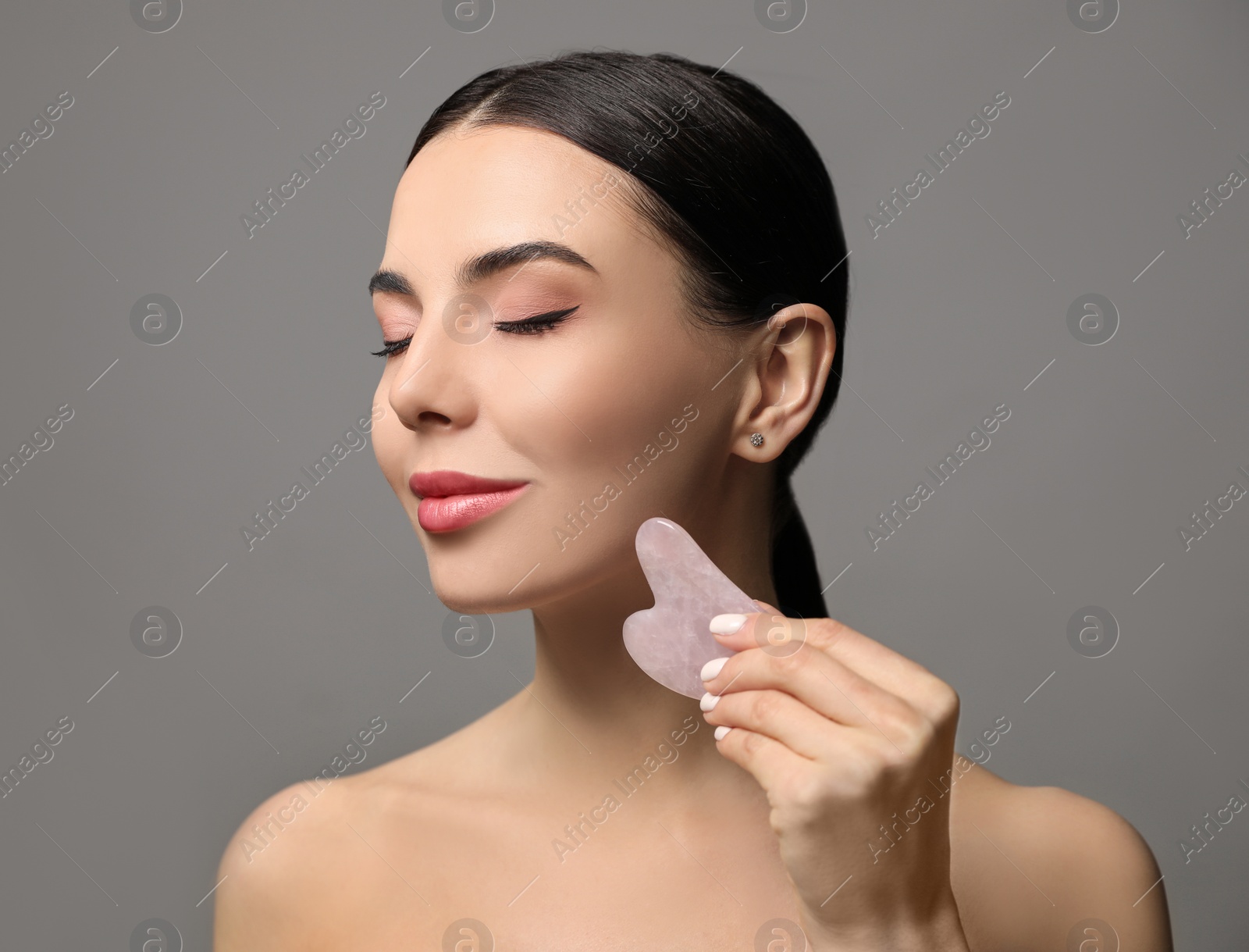 Photo of Beautiful young woman doing facial massage with gua sha tool on grey background