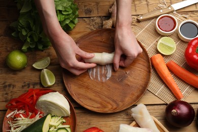 Photo of Making delicious spring rolls. Woman wrapping fresh vegetables into rice paper at wooden table, flat lay