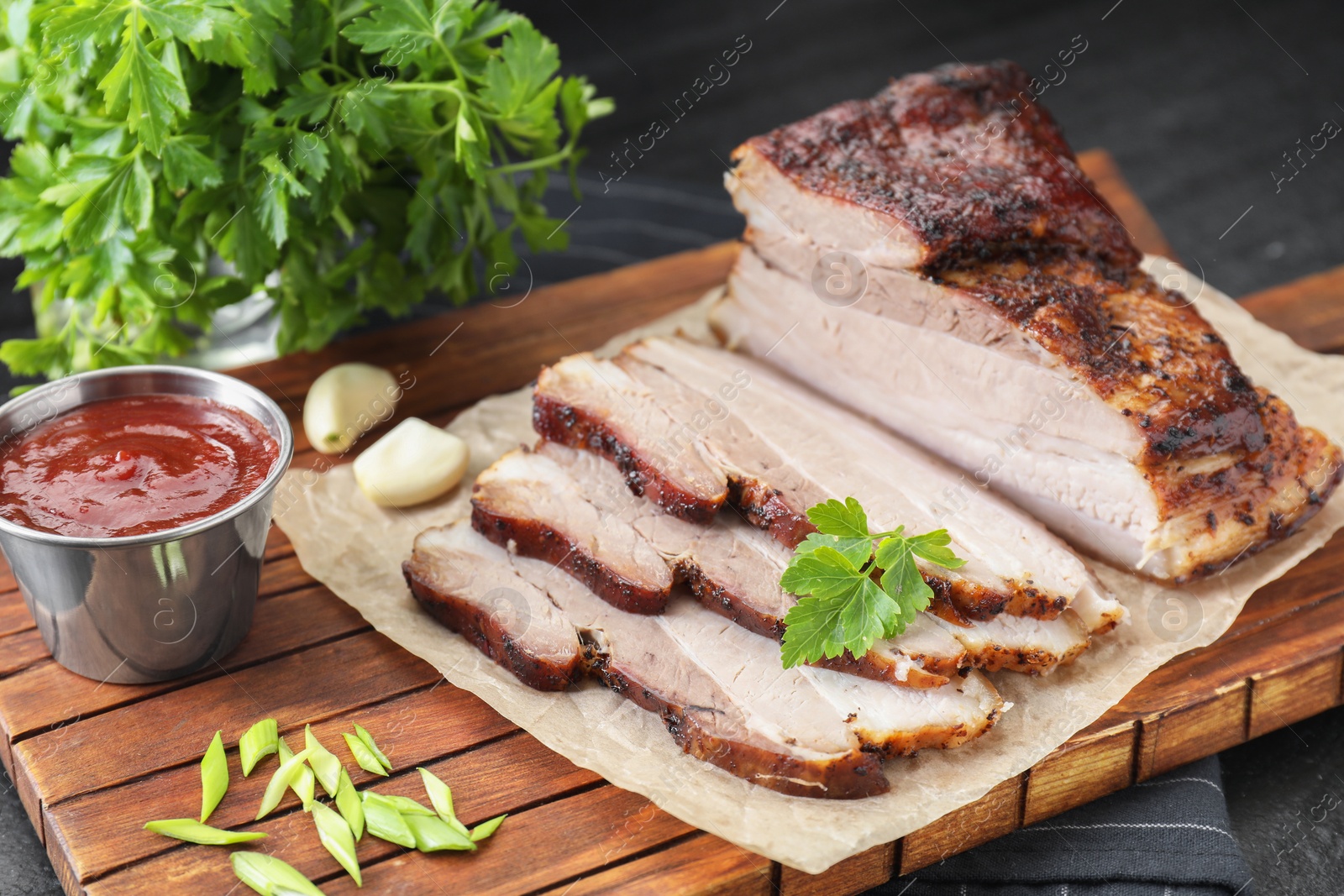 Photo of Pieces of baked pork belly served with sauce and parsley on black textured table, closeup