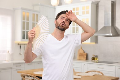 Bearded man waving white hand fan to cool himself in kitchen