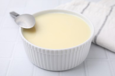 Photo of Bowl with condensed milk and spoon on white tiled table, closeup