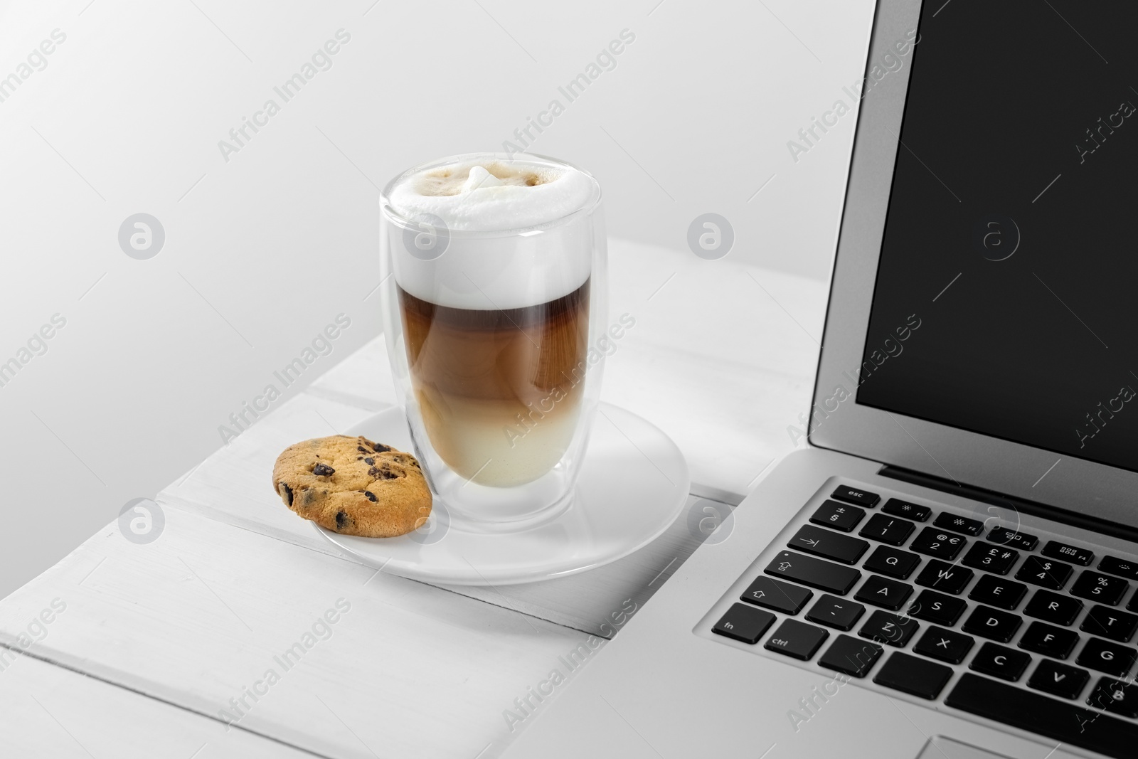 Photo of Chocolate chip cookie, latte and laptop on white wooden table indoors