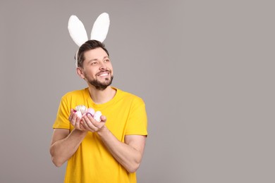 Happy man in bunny ears headband holding painted Easter eggs on grey background. Space for text
