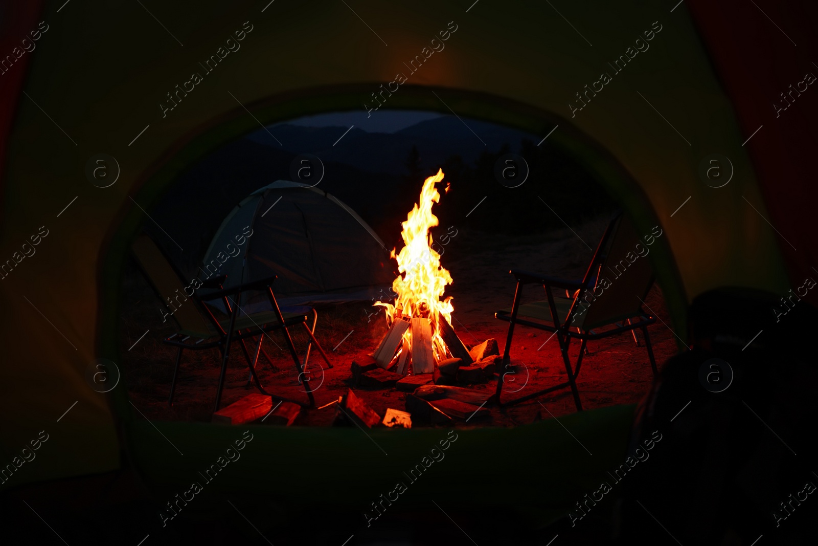 Photo of Beautiful bonfire and folding chairs outdoors in evening, view from camping tent