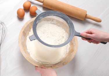 Photo of Woman sieving flour into bowl at white wooden table, above view