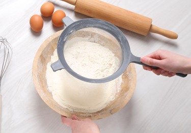 Woman sieving flour into bowl at white wooden table, above view