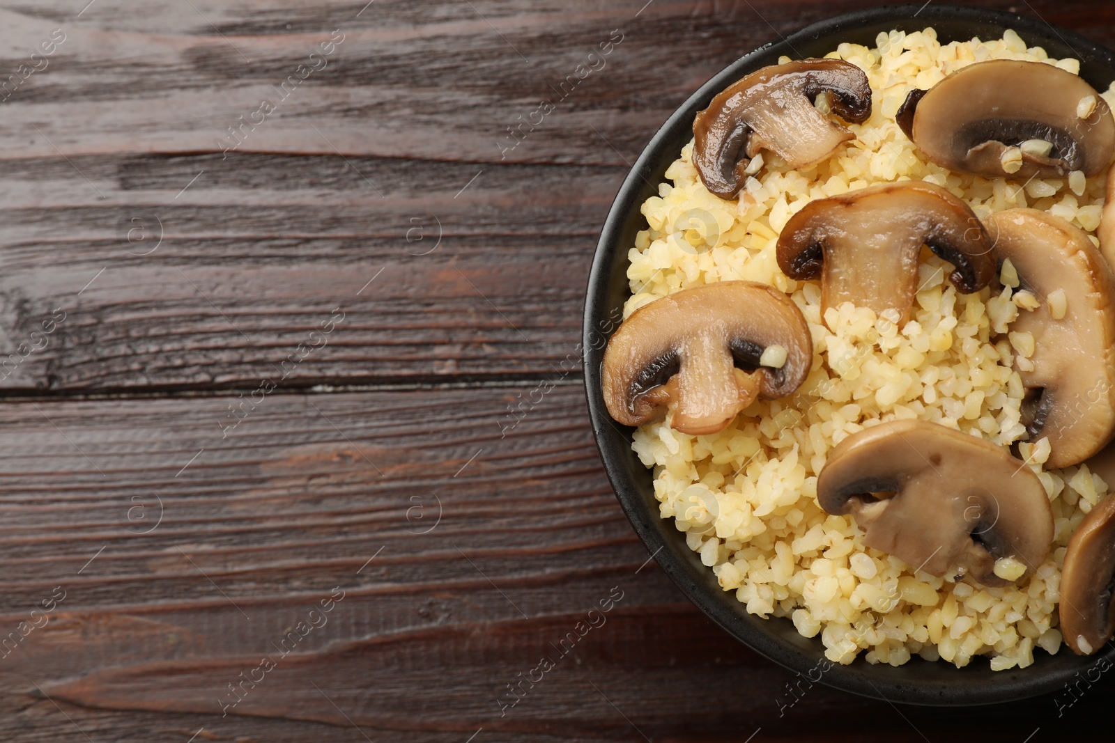 Photo of Delicious bulgur with mushrooms in bowl on wooden table, top view. Space for text