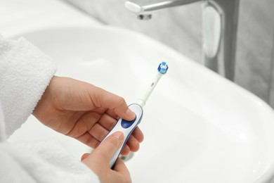 Photo of Woman turning on electric toothbrush near sink in bathroom, closeup