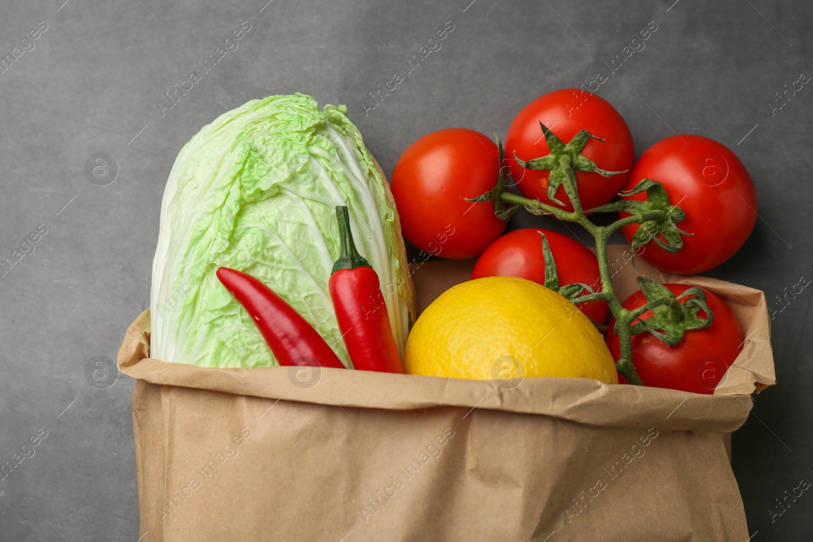 Photo of Paper bag with fresh Chinese cabbage, lemon, tomatoes and chili pepper on grey textured table, flat lay