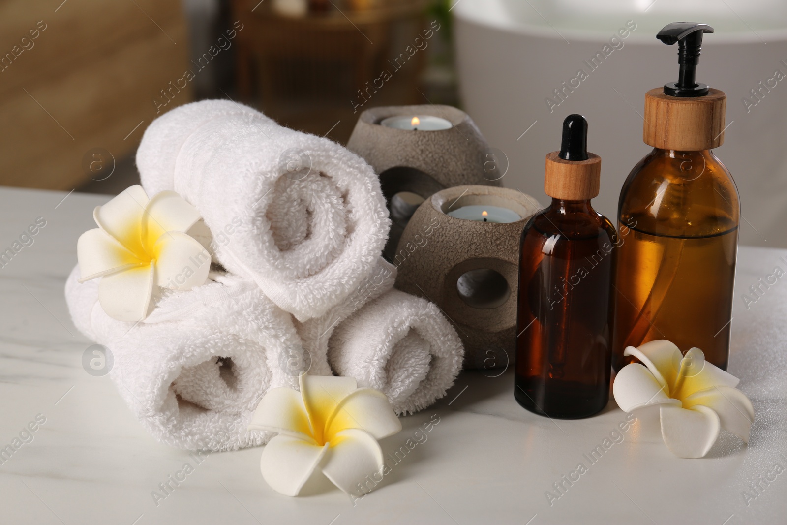 Photo of Spa products, burning candles, towels and plumeria flowers on white table in bathroom, closeup