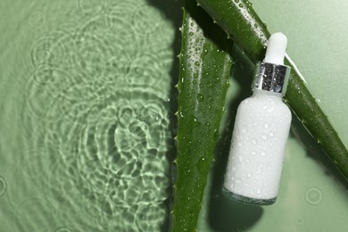 Photo of Bottle of cosmetic product and aloe leaves in water on pale green background, flat lay. Space for text