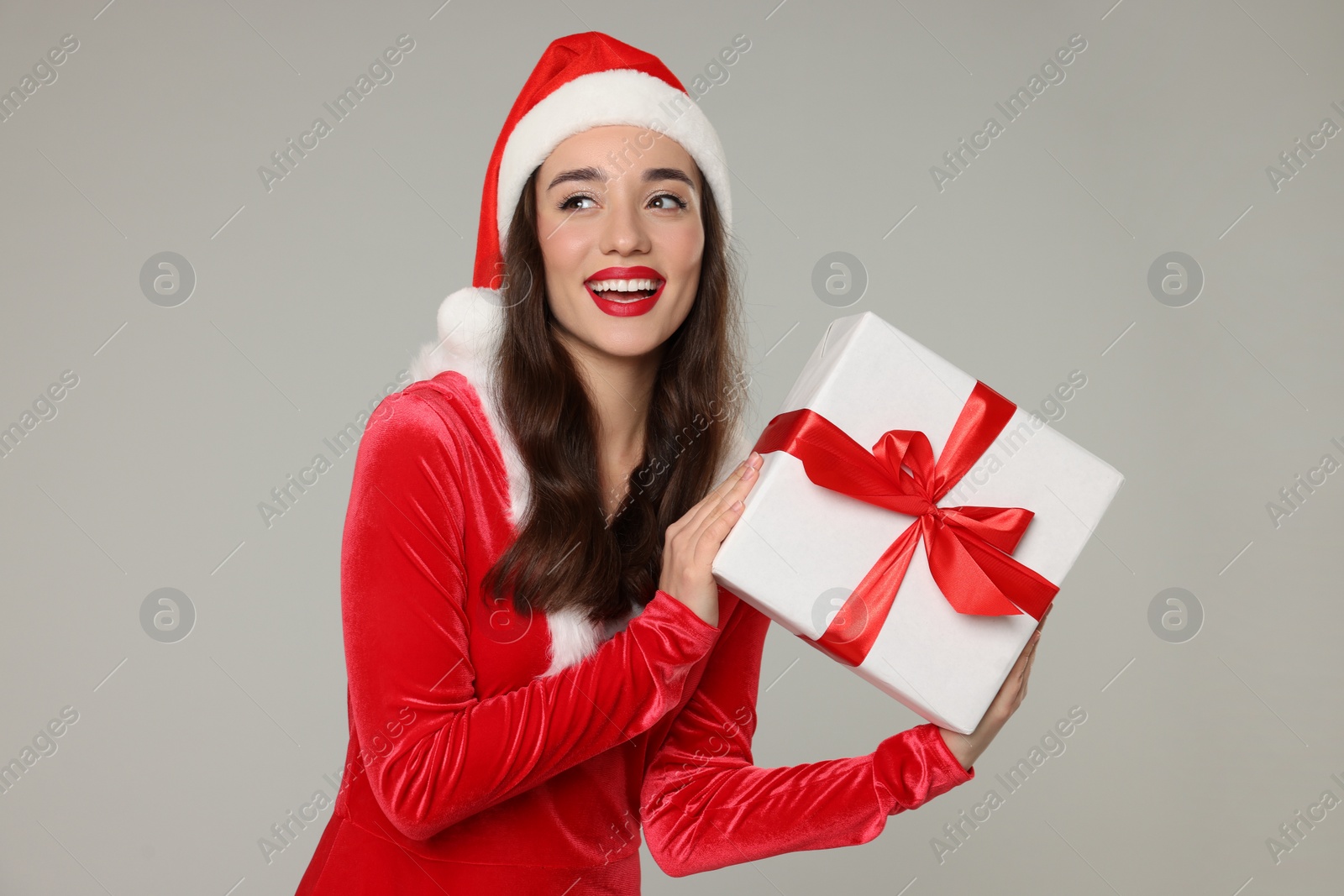 Photo of Beautiful young woman in red dress and Santa hat with Christmas gift on grey background