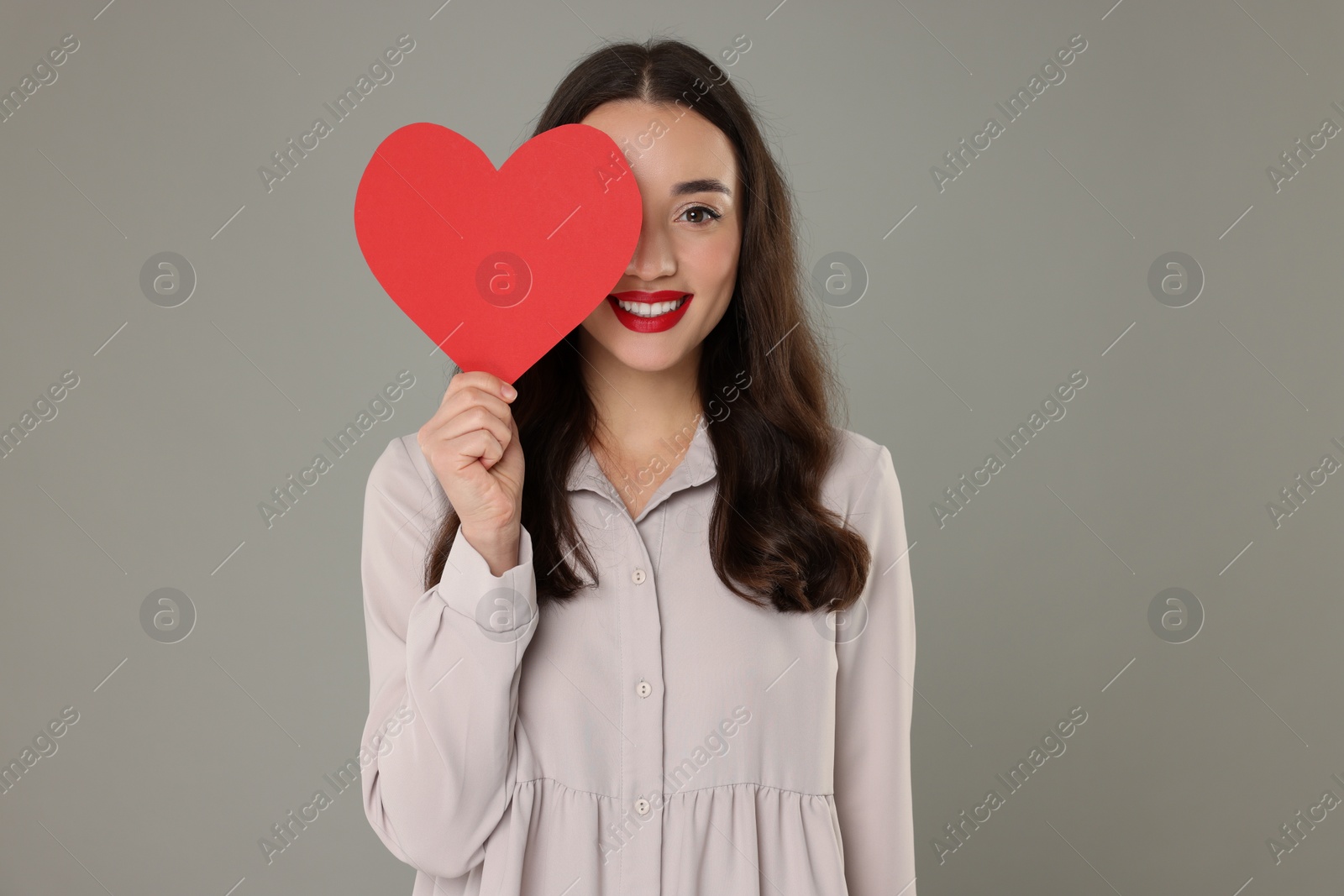 Photo of Beautiful young woman covering her eye with paper heart on grey background