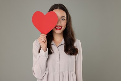 Beautiful young woman covering her eye with paper heart on grey background