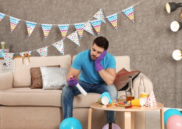 Tired young man with bottle of detergent in messy room after party