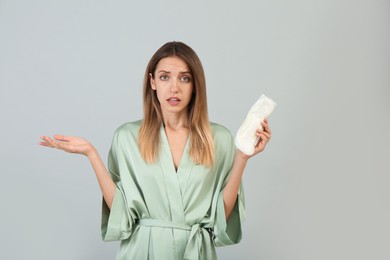 Confused young woman with disposable menstrual pad on grey background