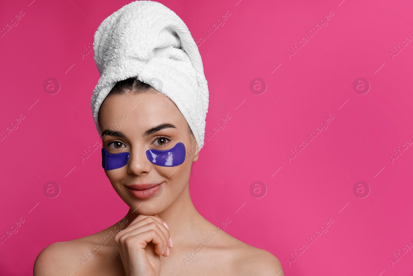 Photo of Beautiful young woman with under eye patches and hair wrapped in towel on pink background, space for text