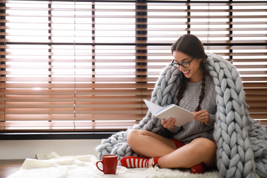 Image of Happy young woman wrapped in knitted blanket with book near window at home. Lazy morning