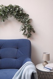 Photo of Beautiful garland made of eucalyptus branches hanging above armchair on white wall indoors