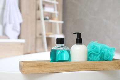 Wooden bath tray with bottles of shower gels and mesh pouf on tub indoors