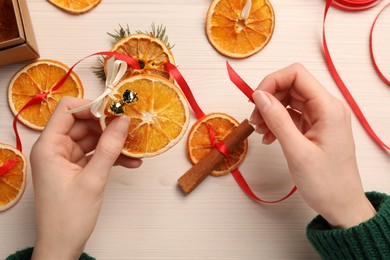 Photo of Woman making garland from dry oranges, cinnamon, ribbon and sleigh bells at white wooden table, top view