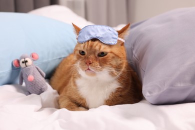 Photo of Cute ginger cat with sleep mask and crocheted mouse resting on bed