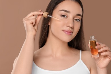 Photo of Beautiful woman applying essential oil onto face on brown background