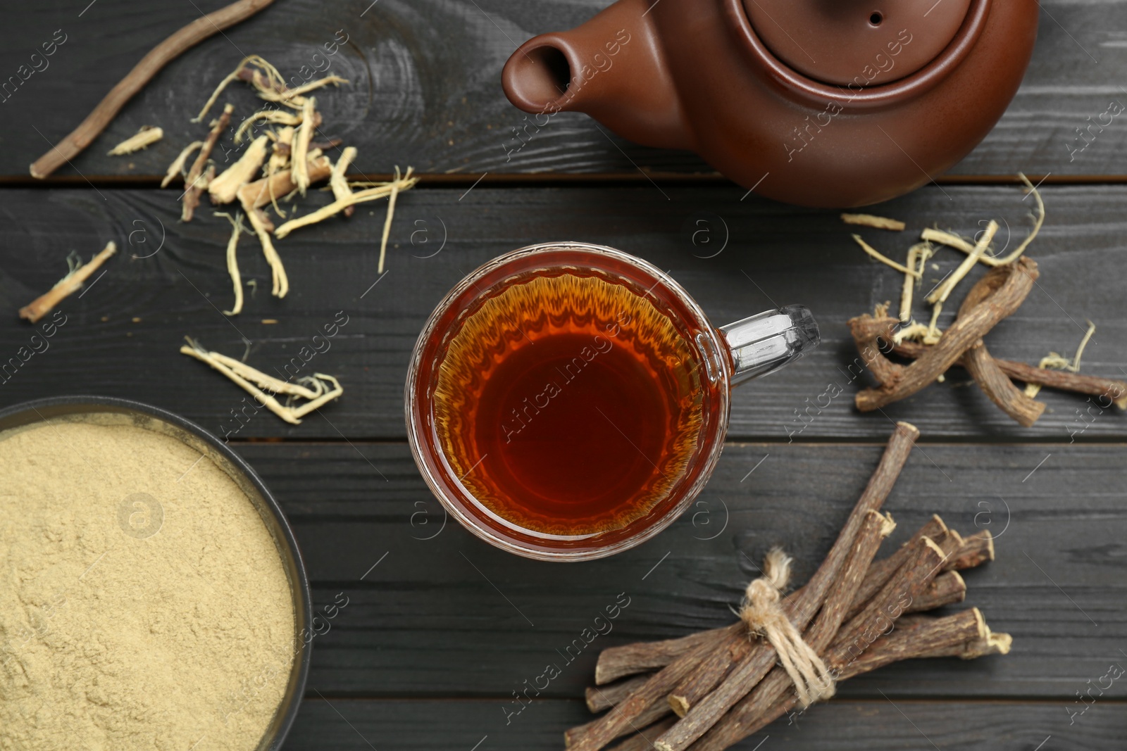 Photo of Aromatic licorice tea in cup, teapot, dried sticks of licorice root and powder on black wooden table, flat lay